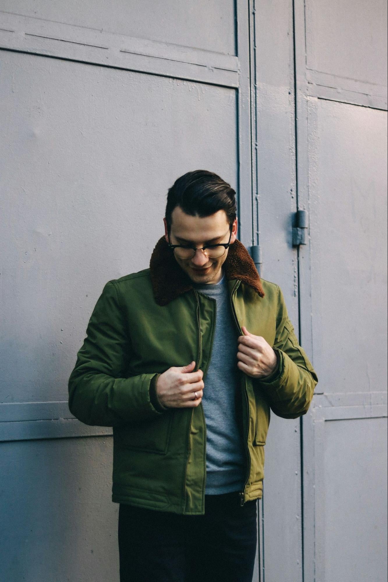 A man in a bomber jacket stands outside of a building.