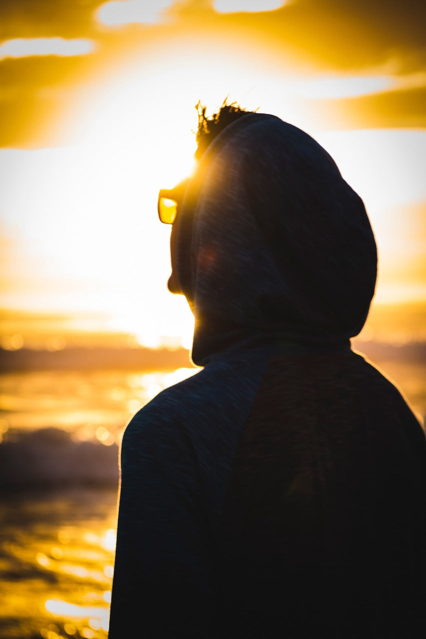 A man wearing a hoodie and sunglasses is at a beach on a sunny day.