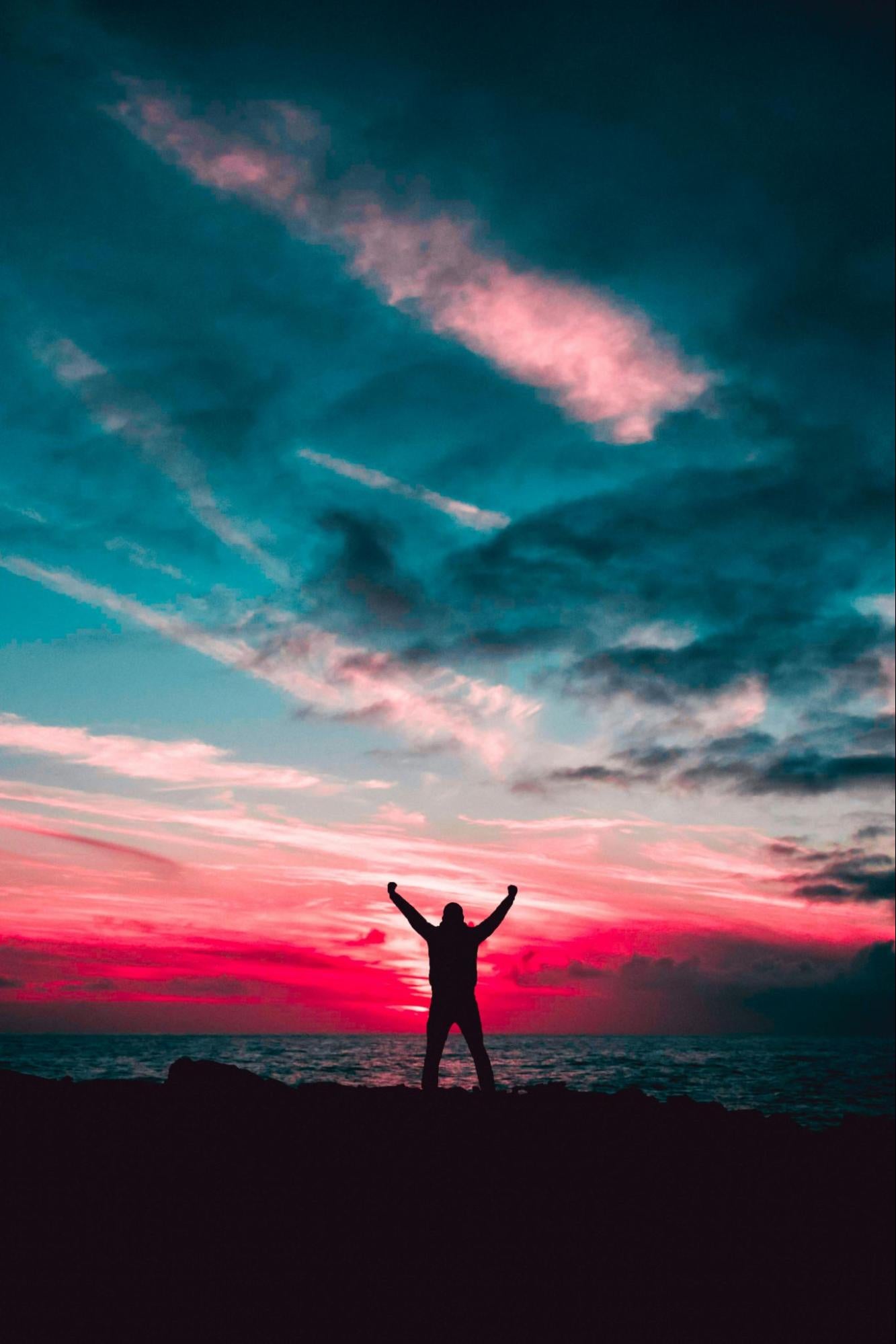 A man with raised arms watches a sunset over the ocean.