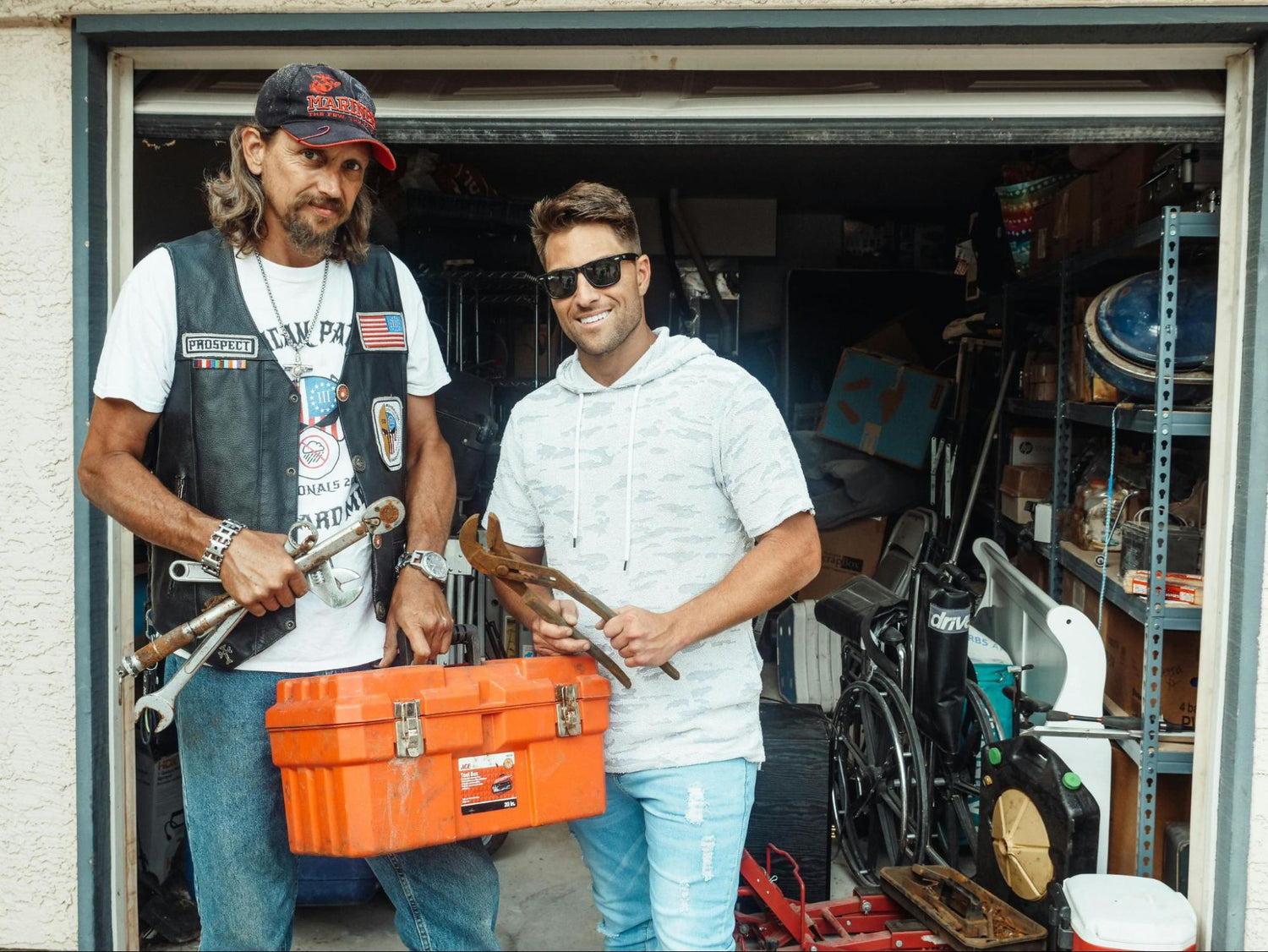 Two men hold tools and a toolbox in front of a cluttered garage.